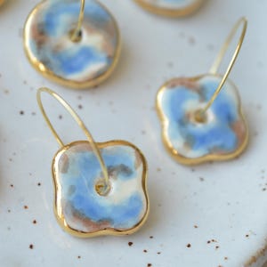 Può includere: Orecchini in ceramica fatti a mano con bordo placcato oro. Gli orecchini presentano un design unico con motivi ad acquerello blu, bianchi e marroni. Gli orecchini sono su cerchi dorati.