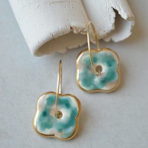 Puede incluir: Un par de pendientes de aro con colgantes cuadrados de bordes redondeados. Los colgantes tienen una base blanca con patrones de acuarela en color verde azulado y rosa suave, con un borde de metal dorado y un aro de alambre fino dorado.