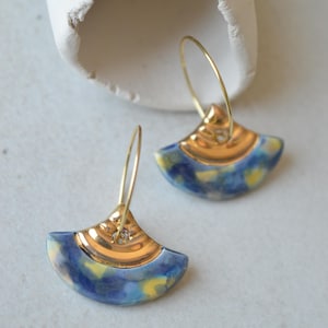 Pode incluir: Um par de brincos em forma de leque com uma secção superior em metal dourado polido e uma base de cerâmica curva com um padrão manchado em azul e amarelo. Cada brinco está suspenso por um fino aro de arame dourado no centro.