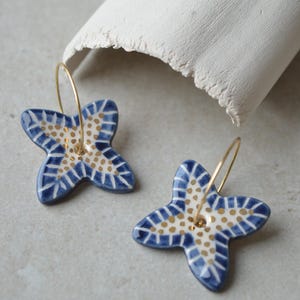 Puede incluir: Un par de pendientes de cerámica azul y blanca con forma de flor y detalles dorados. Los pendientes tienen un diseño de cuatro pétalos y están suspendidos de aros dorados. Los pendientes están sobre una superficie gris claro.