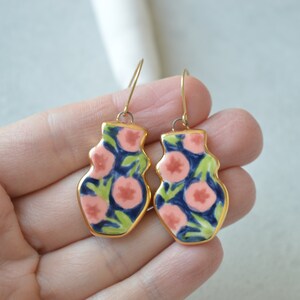 May include: A pair of gold-toned earrings with a floral design. The earrings are shaped like vases and feature pink flowers on a blue and green background.
