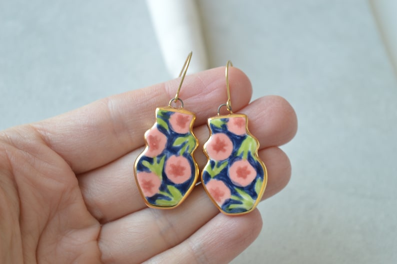 May include: A pair of gold-toned earrings with a floral design. The earrings are shaped like vases and feature pink flowers on a blue and green background.