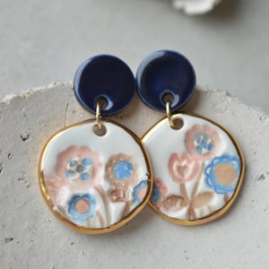 Può includere: Un paio di orecchini pendenti con un perno circolare blu scuro collegato a un ciondolo circolare bianco più grande. Il ciondolo presenta un motivo floreale dipinto a mano nei toni del rosa, blu e marrone chiaro, con un bordo oro metallizzato.