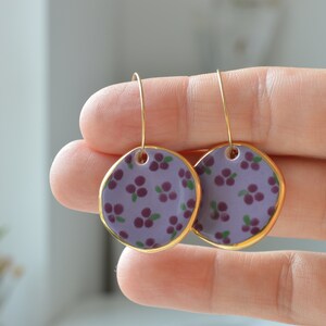 Pode incluir: Um par de brincos de argola dourados com pingentes de cerâmica roxa. Os pingentes são decorados com um pequeno padrão floral em tons de roxo e verde.