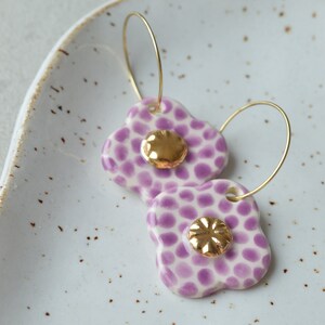 Könnte beinhalten: Ein Paar Ohrringe aus Keramik in Weiß und Lila mit goldenen Akzenten. Die Ohrringe sind blumenförmig mit einem gepunkteten Muster.