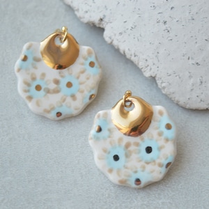 May include: A pair of gold and white ceramic earrings with a floral pattern. The earrings are shaped like a circle with a gold accent on top.