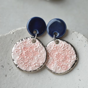 Puede incluir: Un par de pendientes colgantes con un pequeño botón circular azul oscuro brillante conectado a un colgante circular más grande, texturizado y de color rosa pálido. Los colgantes rosas tienen un patrón floral en relieve y un borde plateado.