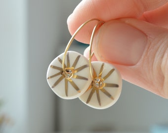 May include: A pair of gold hoop earrings with white ceramic discs featuring a gold starburst design.