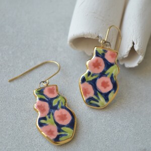 May include: A pair of gold dangle earrings with a floral design. The earrings are made of ceramic and feature pink and green flowers on a dark blue background.