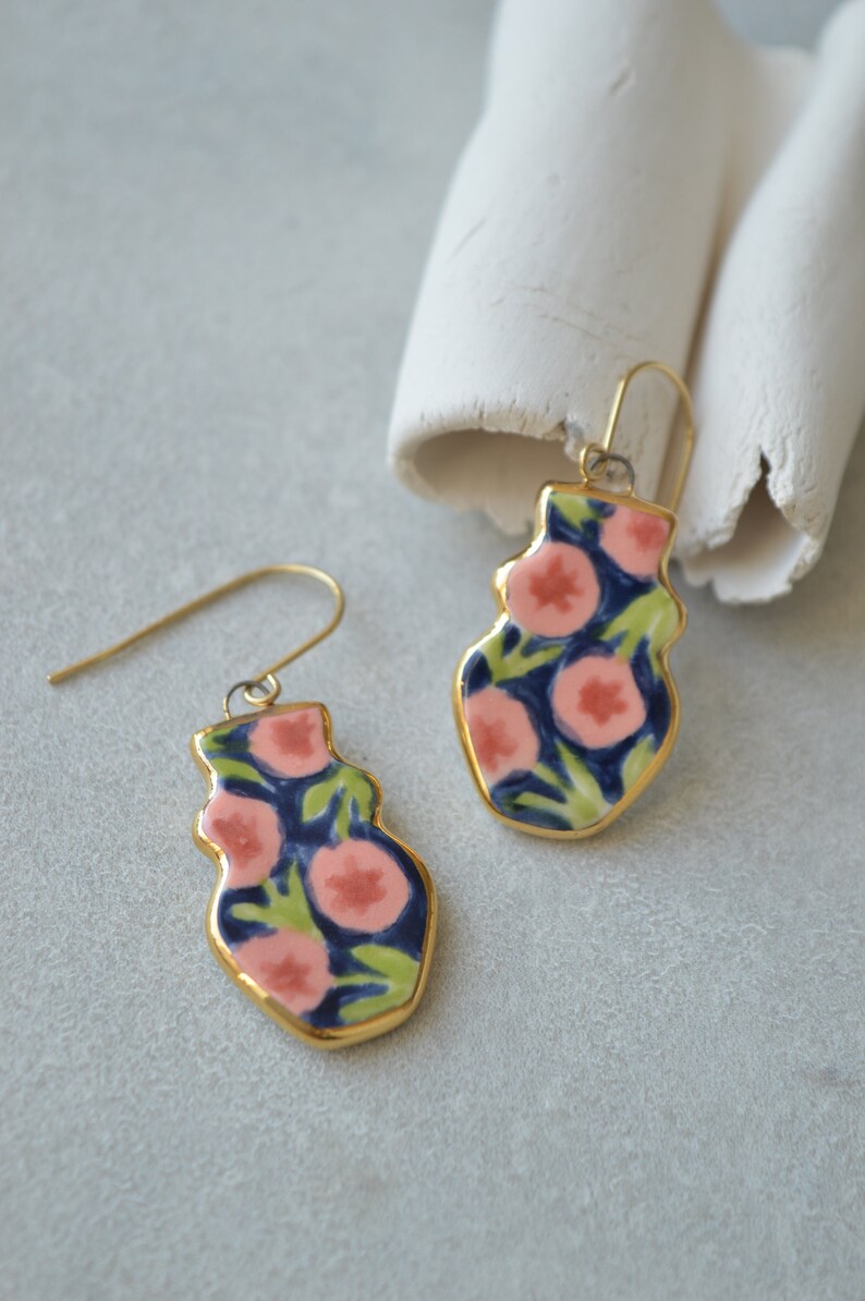 May include: A pair of gold dangle earrings with a floral design. The earrings are made of ceramic and feature pink and green flowers on a dark blue background.