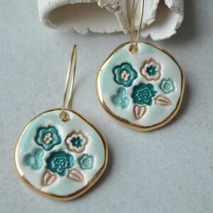 Puede incluir: Un par de pendientes de cerámica con diseño floral. Los pendientes tienen un fondo azul claro con flores en relieve en color turquesa y marrón claro. Los bordes están ribeteados en dorado. Cuelgan de aros dorados.