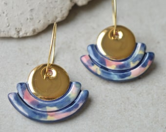 May include: A pair of gold and blue ceramic earrings with a geometric design. The earrings have a gold circle in the center and three blue arches around it. The arches have a pink and white marbled pattern.