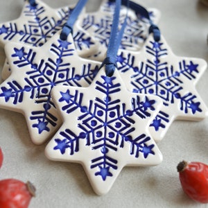 Op de afbeelding: Witte keramische stervormige ornamenten met blauwe sneeuwvlokontwerpen en blauwe linten. De ornamenten zijn gegroepeerd, met rode bessen en eikels op de achtergrond. Perfect voor feestelijke decoratie.