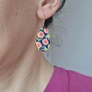 May include: A pair of gold dangle earrings with a pink and blue floral pattern. The earrings are shaped like a vase.