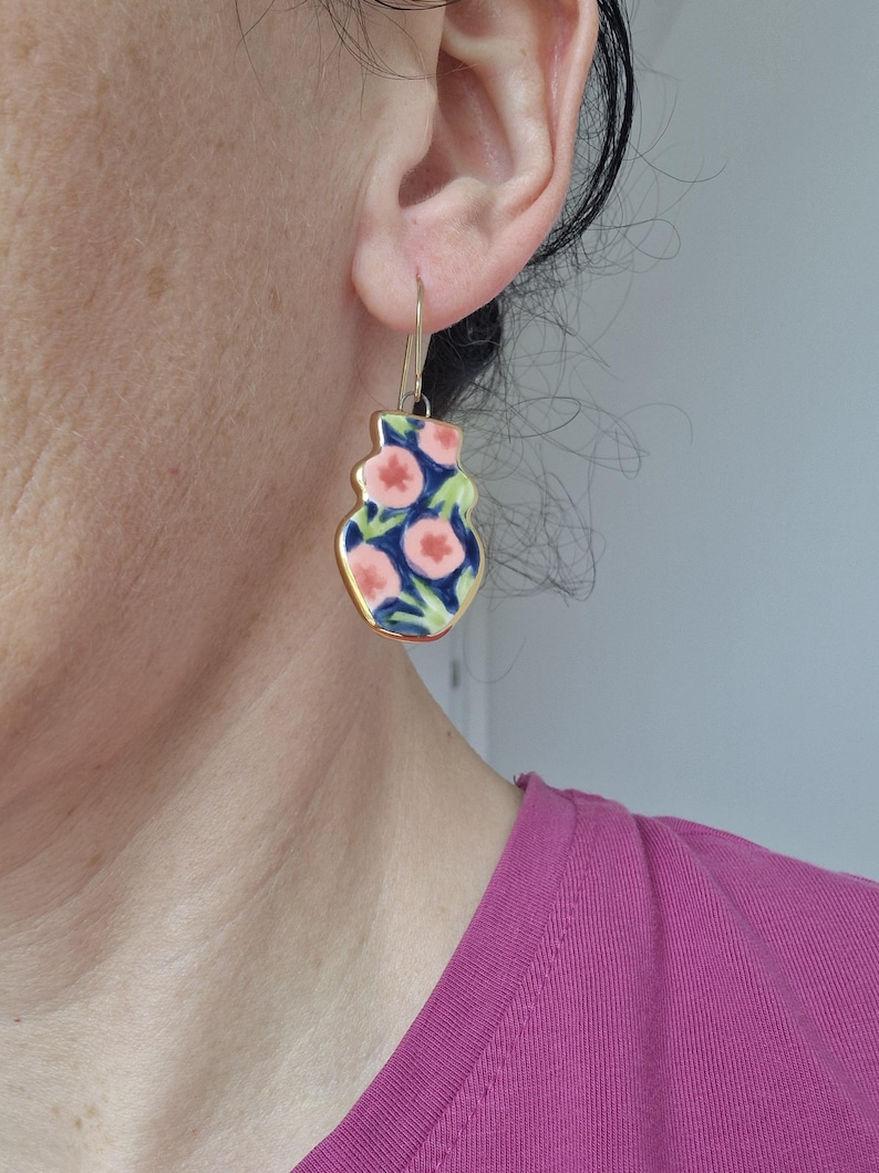 May include: A pair of gold dangle earrings with a pink and blue floral pattern. The earrings are shaped like a vase.