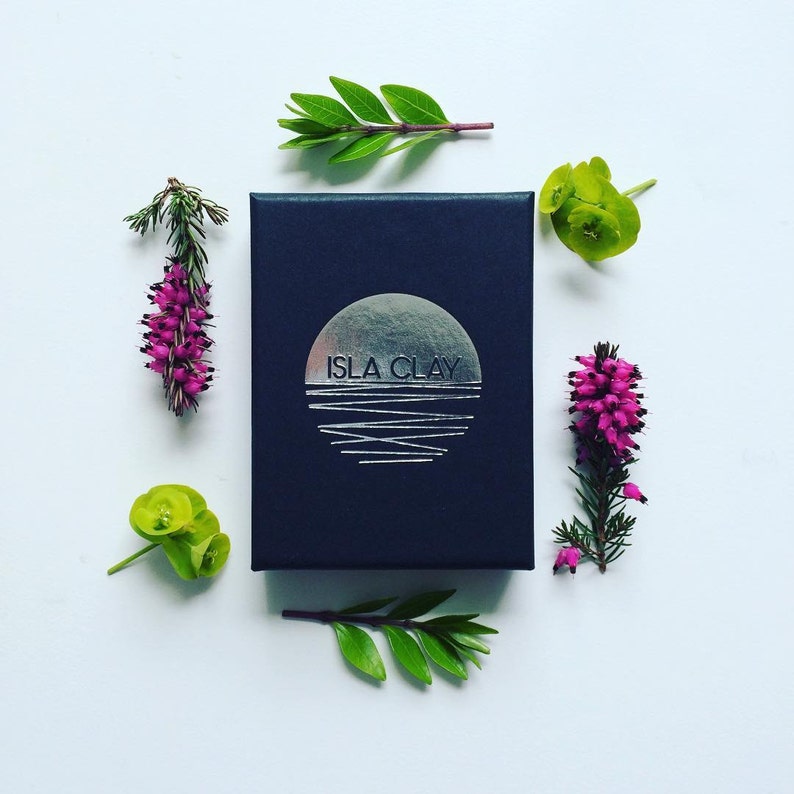 May include: A black box with a silver logo that says "ISLA CLAY" and a silver graphic of a sun setting over water. The box is surrounded by green and pink flowers.