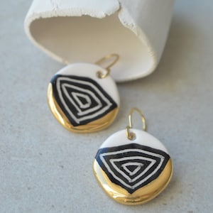 May include: White and gold earrings with a black geometric design. The earrings have a circular shape with a gold-colored bottom half and a white top half. The black design is a series of concentric lines.