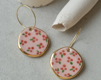 May include: A pair of pink ceramic earrings with a gold rim and a floral pattern. The earrings are on a white surface.