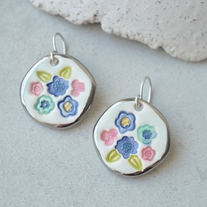 Può includere: Un paio di orecchini in ceramica con un design floreale. Ogni orecchino presenta una base bianca di forma irregolare con un bordo argentato. Il design floreale include fiori blu, rosa e verdi con centri gialli.