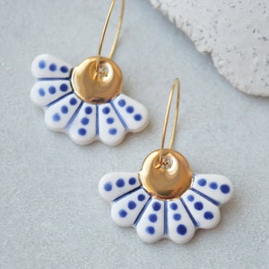 May include: A pair of ceramic earrings featuring a white and blue floral design with gold-colored accents. The earrings have a fan-like shape with blue polka dots and gold-colored hoops.