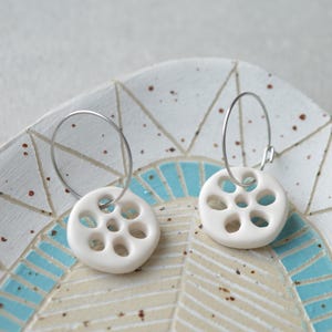 May include: A pair of white ceramic hoop earrings with a circular design featuring multiple cutouts. The earrings are displayed on a patterned ceramic plate.