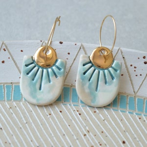 Può includere: Un paio di orecchini in ceramica con una smaltatura blu e bianca e dettagli dorati. Gli orecchini hanno la forma di un semicerchio con un design a raggiera.