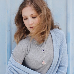 May include: A woman wearing a light blue cardigan over a grey t-shirt. She is wearing two necklaces with heart-shaped pendants. One pendant is blue and white, and the other is pink and white.