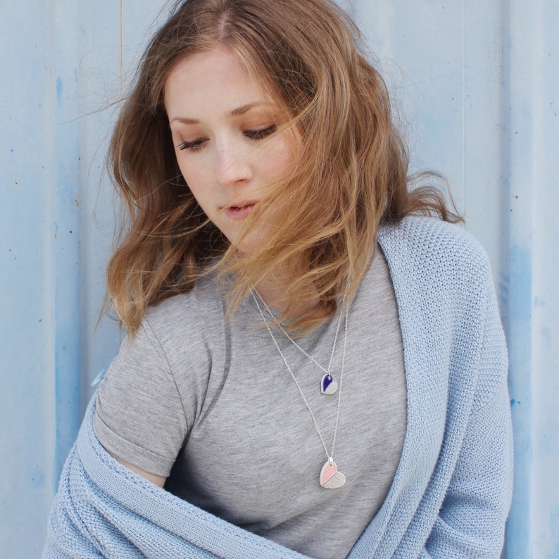 May include: A woman wearing a light blue cardigan over a grey t-shirt. She is wearing two necklaces with heart-shaped pendants. One pendant is blue and white, and the other is pink and white.