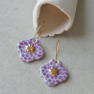 Può includere: Un paio di orecchini a cerchio dorati con ciondoli a forma di fiore in ceramica bianca e viola. I fiori hanno una superficie testurizzata e un centro dorato.