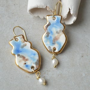Op de afbeelding: Een paar keramische oorbellen met een gouden rand en een unieke vaasvorm. De oorbellen hebben een blauw, wit en bruin aquarelontwerp en een kleine pareldruppel. De oorbellen worden op een neutrale ondergrond gepresenteerd.