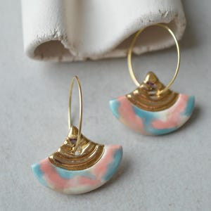 Puede incluir: Un par de pendientes de aro dorados con colgantes en forma de abanico. Los colgantes presentan una combinación de colores pastel rosa, azul y blanco, acentuados con detalles metálicos dorados. Los pendientes se muestran sobre un fondo neutro.
