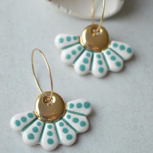 Puede incluir: Un par de pendientes de cerámica blancos y verde azulado con un diseño floral. Cada pendiente presenta un perno circular de color dorado y un colgante en forma de abanico con lunares verde azulado. Los pendientes están en aros dorados.