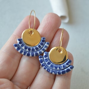 Könnte beinhalten: Ein Paar goldene Creolen-Ohrringe mit einem fächerförmigen Anhänger. Der Anhänger hat eine goldene Scheibe über einem blau-weiß gemusterten Keramikdesign. Die Ohrringe werden vor einem neutralen Hintergrund gezeigt.