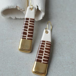 Può includere: Un paio di lunghi orecchini pendenti rettangolari in ceramica con una parte superiore color crema, una sezione centrale con un motivo a griglia marrone e bianco e una base dipinta in oro metallizzato. Sono appesi a sottili ganci dorati.