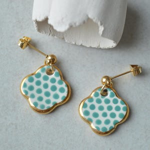 Può includere: Un paio di orecchini color oro con un design a quadrifoglio. I ciondoli a forma di quadrifoglio sono bianchi con pois turchese e un bordo dorato. Gli orecchini hanno un perno e un piccolo borchia dorata.