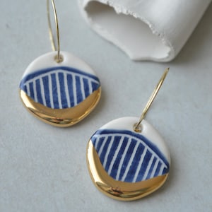 Könnte beinhalten: Ein Paar Keramikohrringe. Jeder Ohrring hat eine weiße, abgerundete Scheibe mit einem blau-weiß gestreiften Design und einem goldfarbenen, gebogenen Boden. Die Ohrringe hängen an goldfarbenen Reifen.