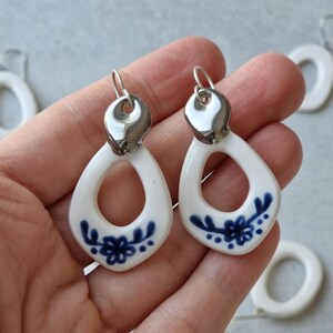 Pode incluir: Um par de brincos de cerâmica branca com um design floral azul. Os brincos têm um gancho prateado e uma forma de lágrima.