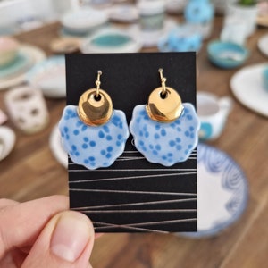 Può includere: Un paio di orecchini in ceramica blu e oro con un design floreale. Gli orecchini hanno un cerchio dorato al centro e un motivo floreale blu attorno al cerchio.