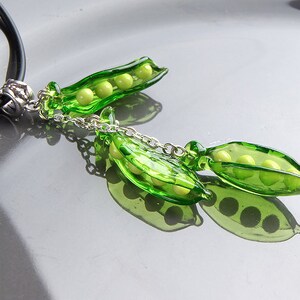 May include: A necklace with a pendant featuring two green glass pea pods with yellow glass peas inside. The pendant is attached to a black cord with a silver clasp.