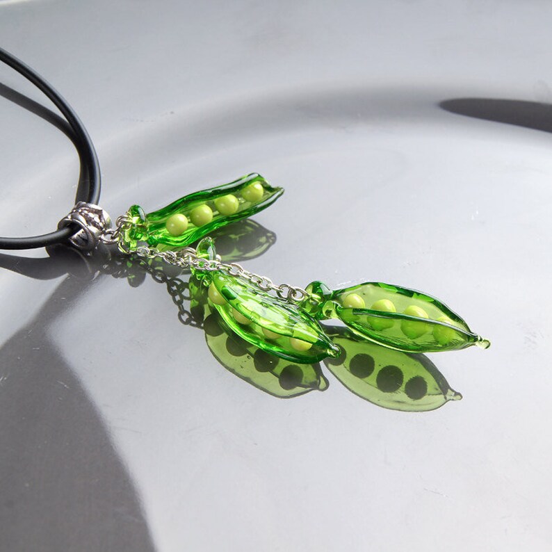May include: A necklace with a pendant featuring two green glass pea pods with yellow glass peas inside. The pendant is attached to a black cord with a silver clasp.