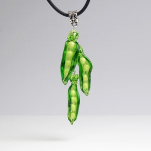 May include: A glass pendant necklace featuring three green pea pods with white pea beads inside. The pendant is attached to a black cord.