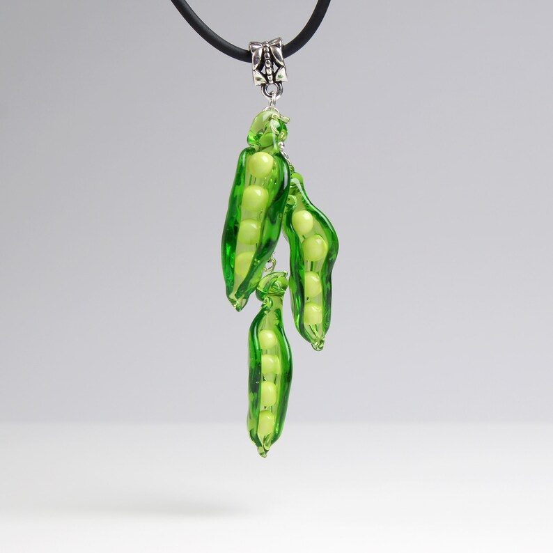 May include: A glass pendant necklace featuring three green pea pods with white pea beads inside. The pendant is attached to a black cord.