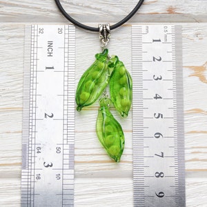 May include: A green glass pea pod pendant necklace with a black cord. The pendant is about 2 inches long and features three pea pods with green glass peas inside. The necklace is measured against a ruler.