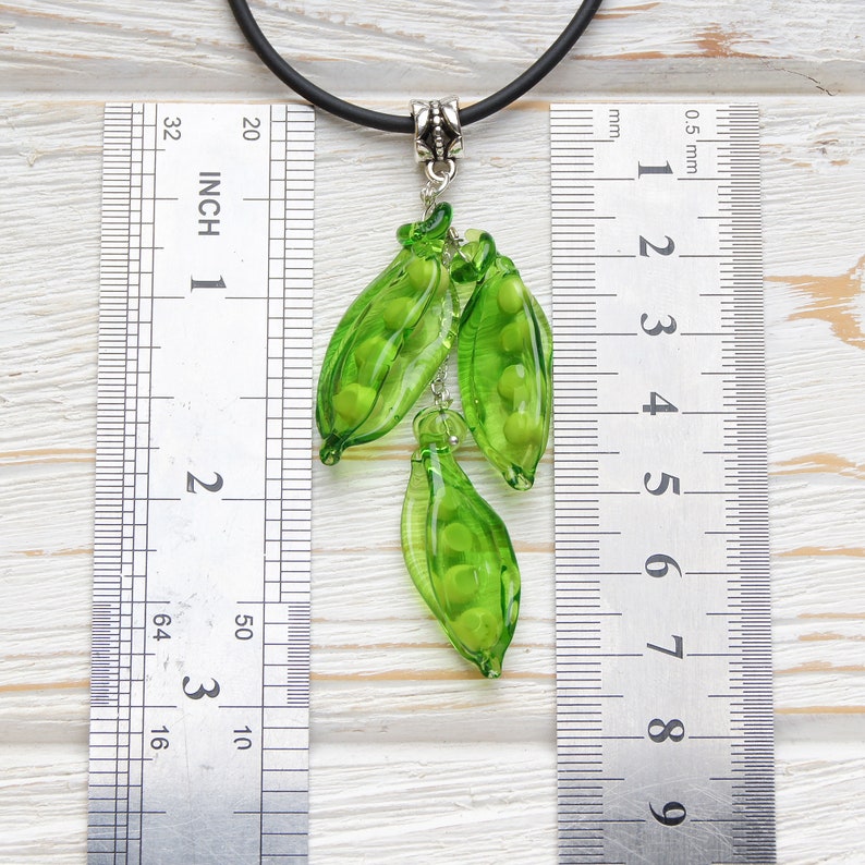 May include: A green glass pea pod pendant necklace with a black cord. The pendant is about 2 inches long and features three pea pods with green glass peas inside. The necklace is measured against a ruler.