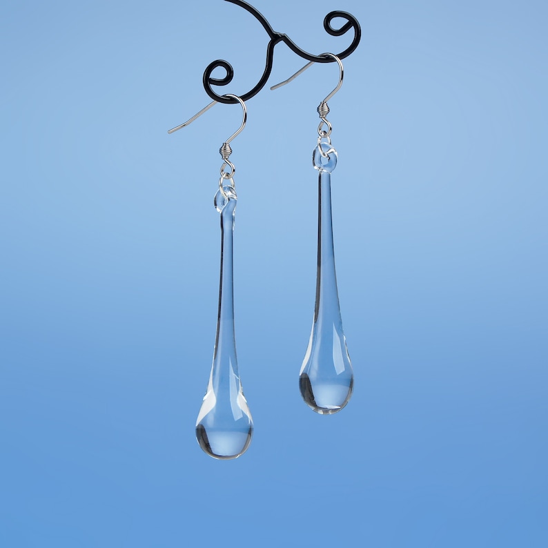 May include: A pair of clear glass teardrop earrings hanging from a silver hook. The earrings are suspended from a black metal earring stand against a light blue background.