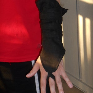 May include: A black fabric arm guard with a long, pointed finger covering. The guard is worn on the left arm and is secured with a wrap around the forearm.
