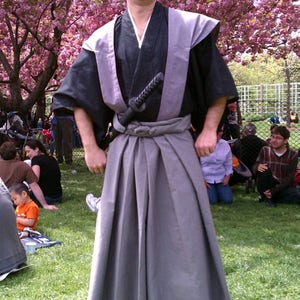 May include: A person wearing a traditional Japanese samurai outfit, including a black kimono, gray hakama pants, and a katana sword. The person is standing in a park with a cherry blossom tree in the background.