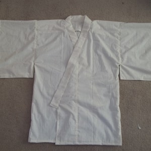 Puede incluir: Un kimono blanco con manga larga y recta. El kimono está hecho de una tela ligera y tiene un diseño sencillo.