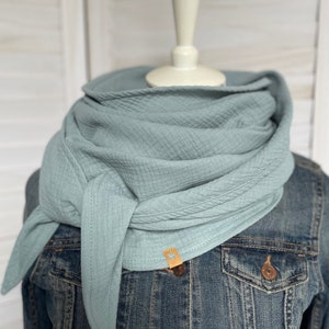 May include: A light blue muslin scarf is draped around a mannequin's neck, knotted at the front. The scarf has a textured appearance and is paired with a denim jacket. A small, tan leather tag is sewn on the scarf.