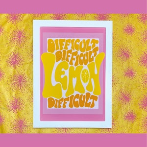 May include: A white framed print with a pink background and the text "Difficult Lemon Difficult" in a retro font. The word "Lemon" is in yellow and orange with a small lemon slice inside the "O".